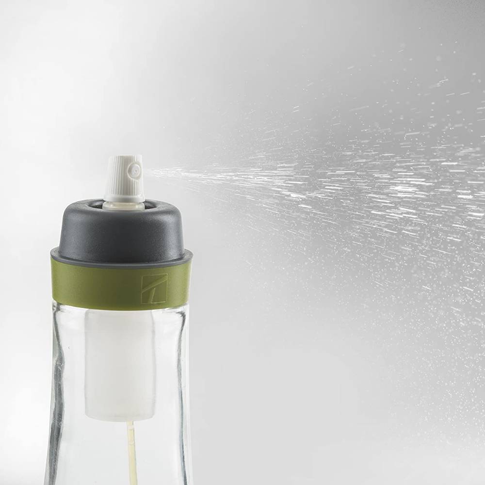 Clear glass oil spray bottle with a green silicone band and white spray nozzle releasing a fine mist, designed for precise oil application in kitchen and cookware use.