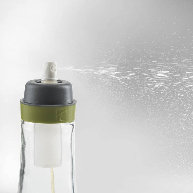 Clear glass oil spray bottle with a green silicone band and white spray nozzle releasing a fine mist, designed for precise oil application in kitchen and cookware use.