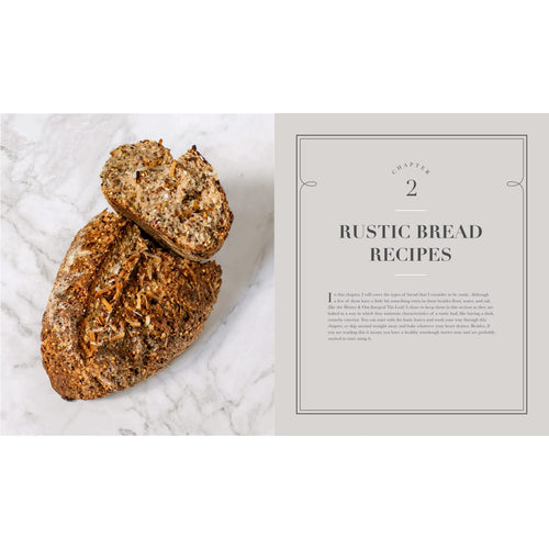 Rustic loaf of multigrain bread with seeds and nuts sliced open next to an open cookbook page titled "Rustic Bread Recipes," suitable for Books, Cookbooks, and Home and Garden How-To Books collections.