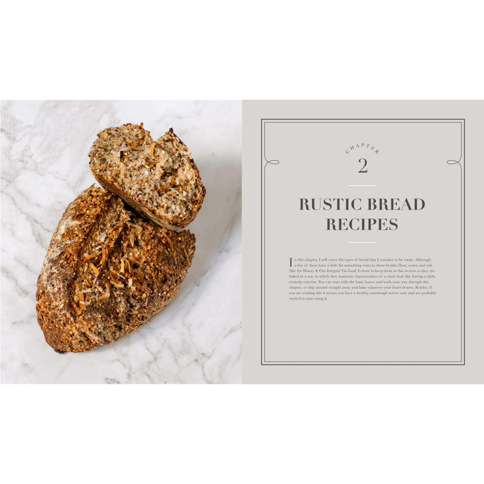 Rustic loaf of multigrain bread with seeds and nuts sliced open next to an open cookbook page titled "Rustic Bread Recipes," suitable for Books, Cookbooks, and Home and Garden How-To Books collections.
