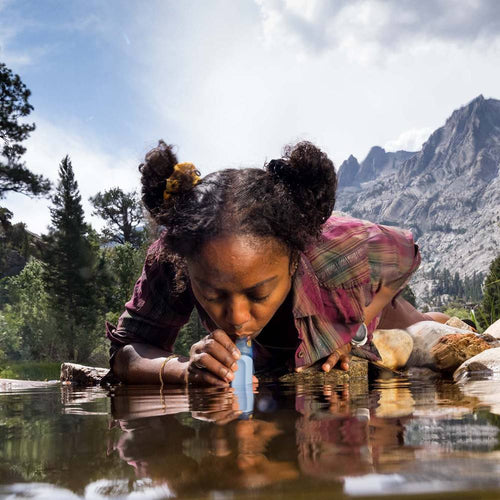 Young woman drinking water directly from a mountain stream using a portable blue water filter straw, highlighting safe hydration for camping and emergency supplies collections.