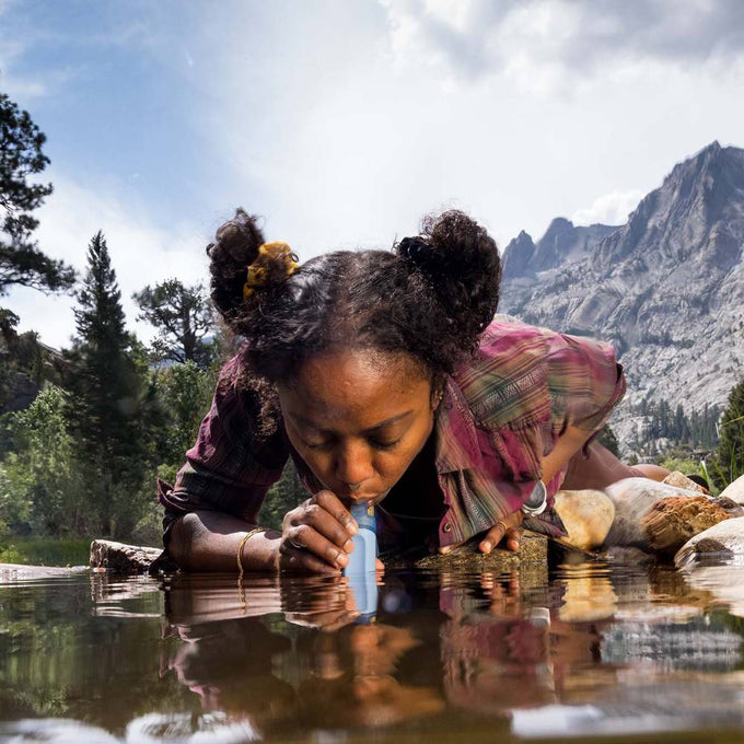 Young woman drinking water directly from a mountain stream using a portable blue water filter straw, highlighting safe hydration for camping and emergency supplies collections.