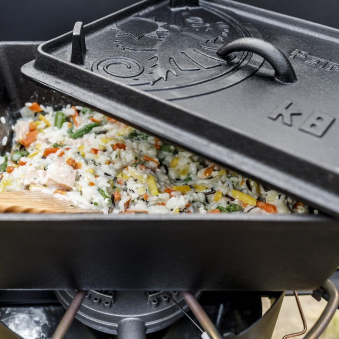 Close-up of a sturdy cast iron loaf pan partially covered with a lid, filled with mixed rice and vegetables, set on a portable camping stove, ideal for outdoor cooking and bread making.