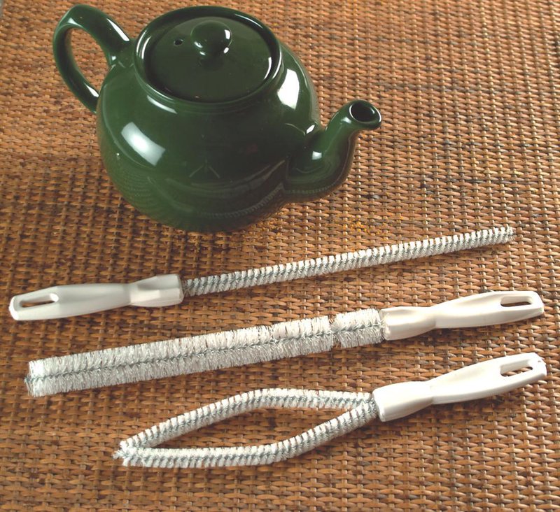 Set of three flexible nylon-bristle brushes with white handles designed for cleaning curved teakettle spouts, displayed next to a dark green ceramic teapot on a woven mat, USA made.