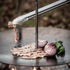 Brushed stainless steel BBQ and coal tongs lifting cooked bacon on a round outdoor griddle with sliced red onions and green peppers, ideal for grilling and camping collections.