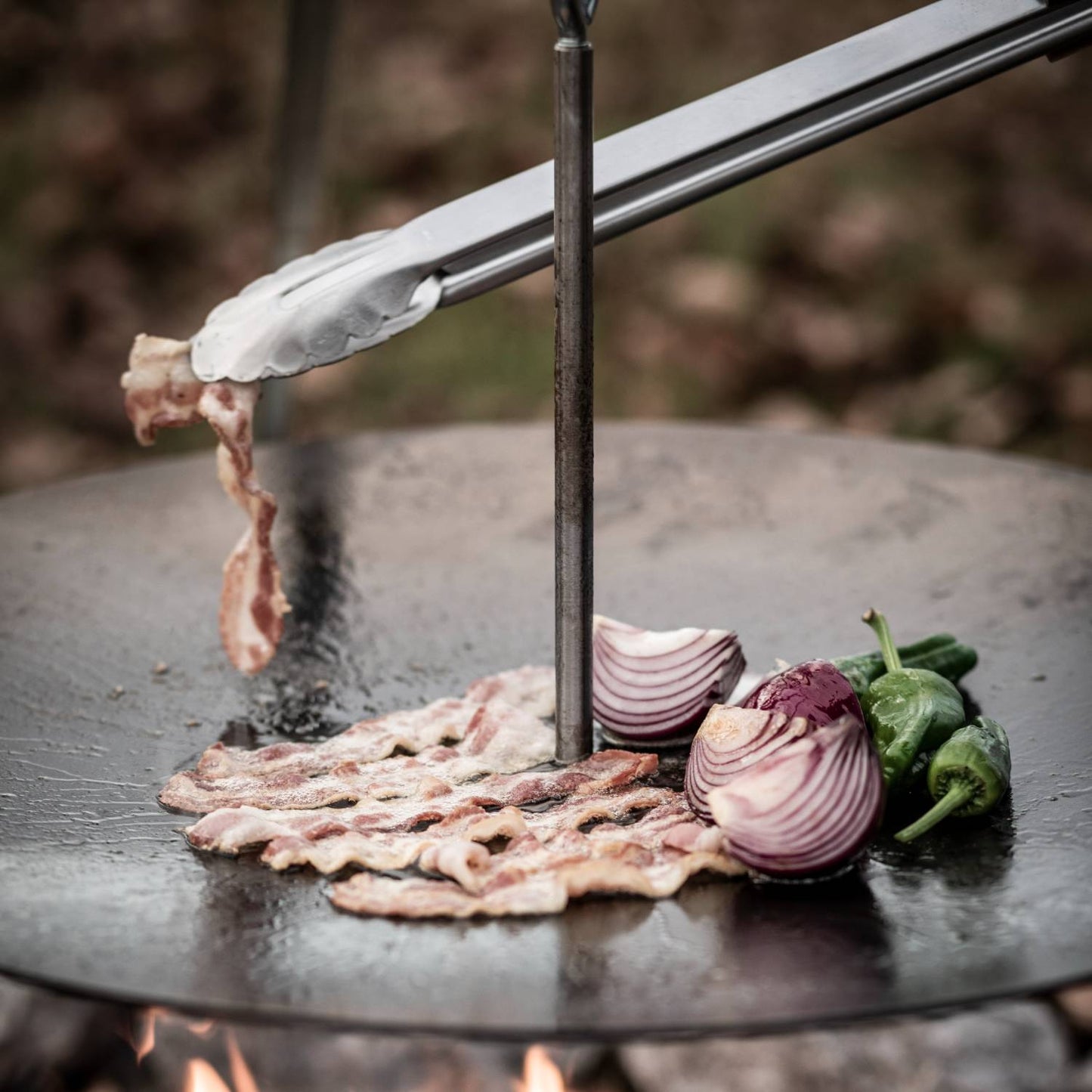 Brushed stainless steel BBQ and coal tongs lifting cooked bacon on a round outdoor griddle with sliced red onions and green peppers, ideal for grilling and camping collections.