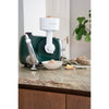 Green stand mixer fitted with a white flake mill attachment rolling grains into flakes collected in a bowl on a granite countertop, surrounded by bread and butter, suited for Kitchen & Food, Baking Supplies, Grain Mills, and Best Sellers collections.