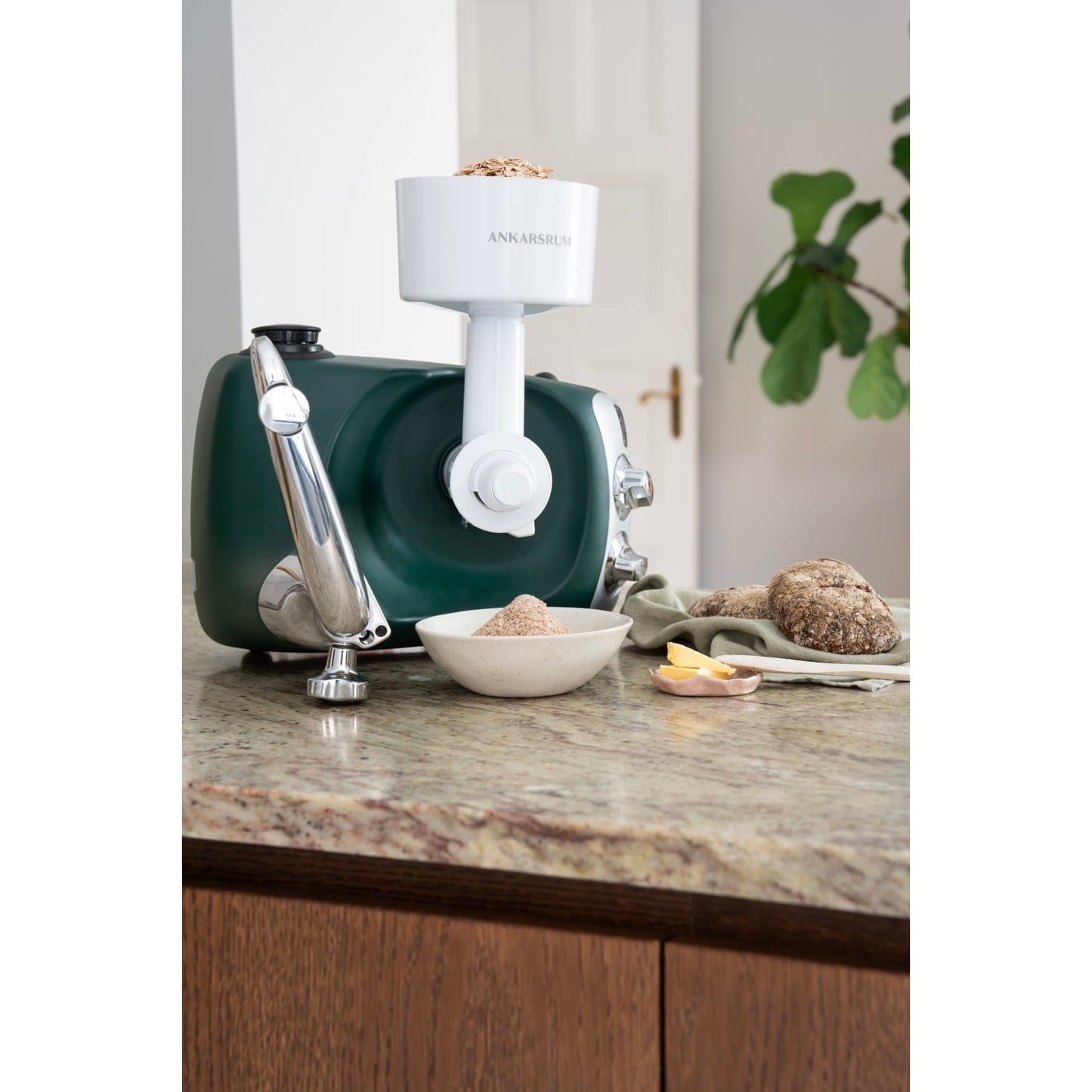 Green stand mixer fitted with a white flake mill attachment rolling grains into flakes collected in a bowl on a granite countertop, surrounded by bread and butter, suited for Kitchen & Food, Baking Supplies, Grain Mills, and Best Sellers collections.