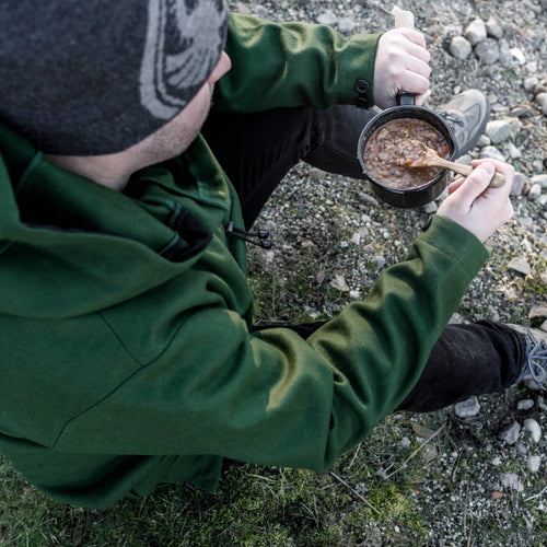Person wearing a green jacket stirring a hearty meal inside a black Petromax enamel pan while seated outdoors on rocky ground, ideal for camping and outdoor cooking collections.