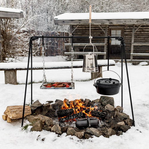 Black coated steel Petromax Fire Bridge with four-leg stability, featuring a hanging grill grate, cast iron pot, and kettle over a stone-bordered campfire in a snowy outdoor setting, ideal for Farm, Garden & Tool, Stoves, Camping, and Grilling collections.