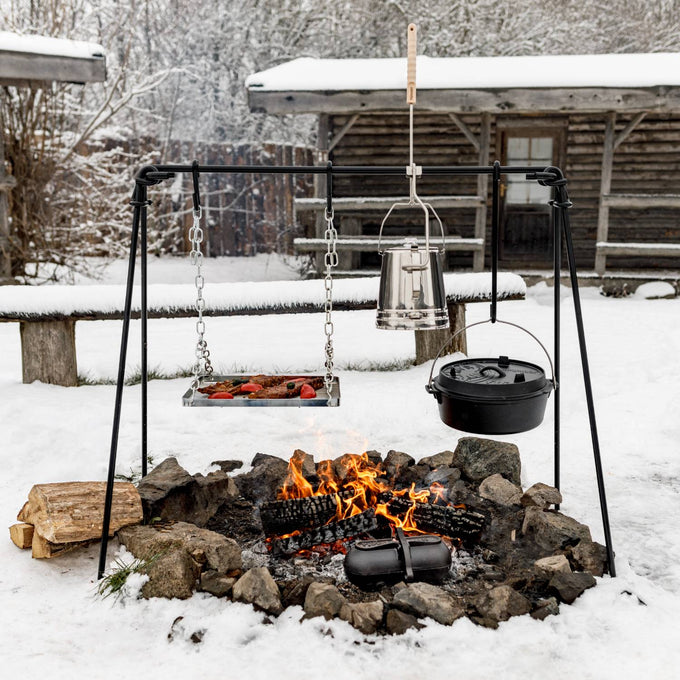Black coated steel Petromax Fire Bridge with four-leg stability, featuring a hanging grill grate, cast iron pot, and kettle over a stone-bordered campfire in a snowy outdoor setting, ideal for Farm, Garden & Tool, Stoves, Camping, and Grilling collections.