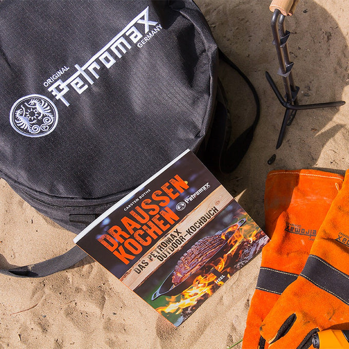 Black durable fabric Petromax transport bag resting on sandy ground next to orange heat-resistant gloves and an outdoor cooking book, suited for Farm, Garden & Tool and Camping collections.