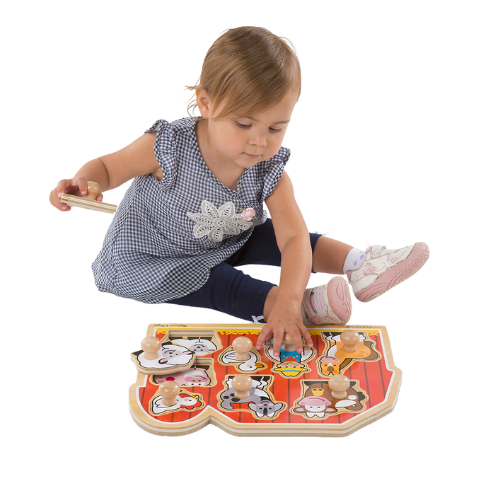 Toddler sitting and playing with a handcrafted wooden farm animal puzzle featuring jumbo knobs on a bright red barn background, encouraging fine motor skills development in children.