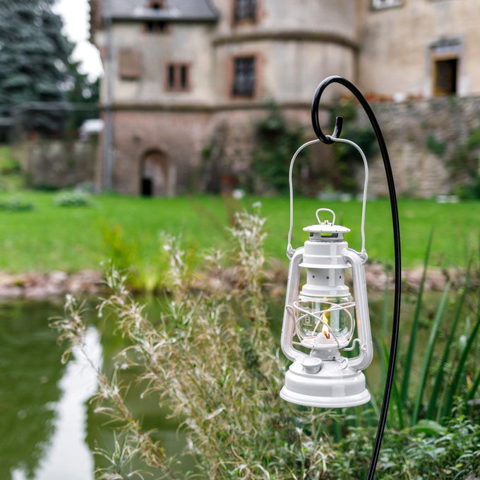 Black metal curved lantern holder suspending a lit white lantern outdoors near a pond with historic building in the background, handcrafted for camping, barn, and emergency supplies collections.