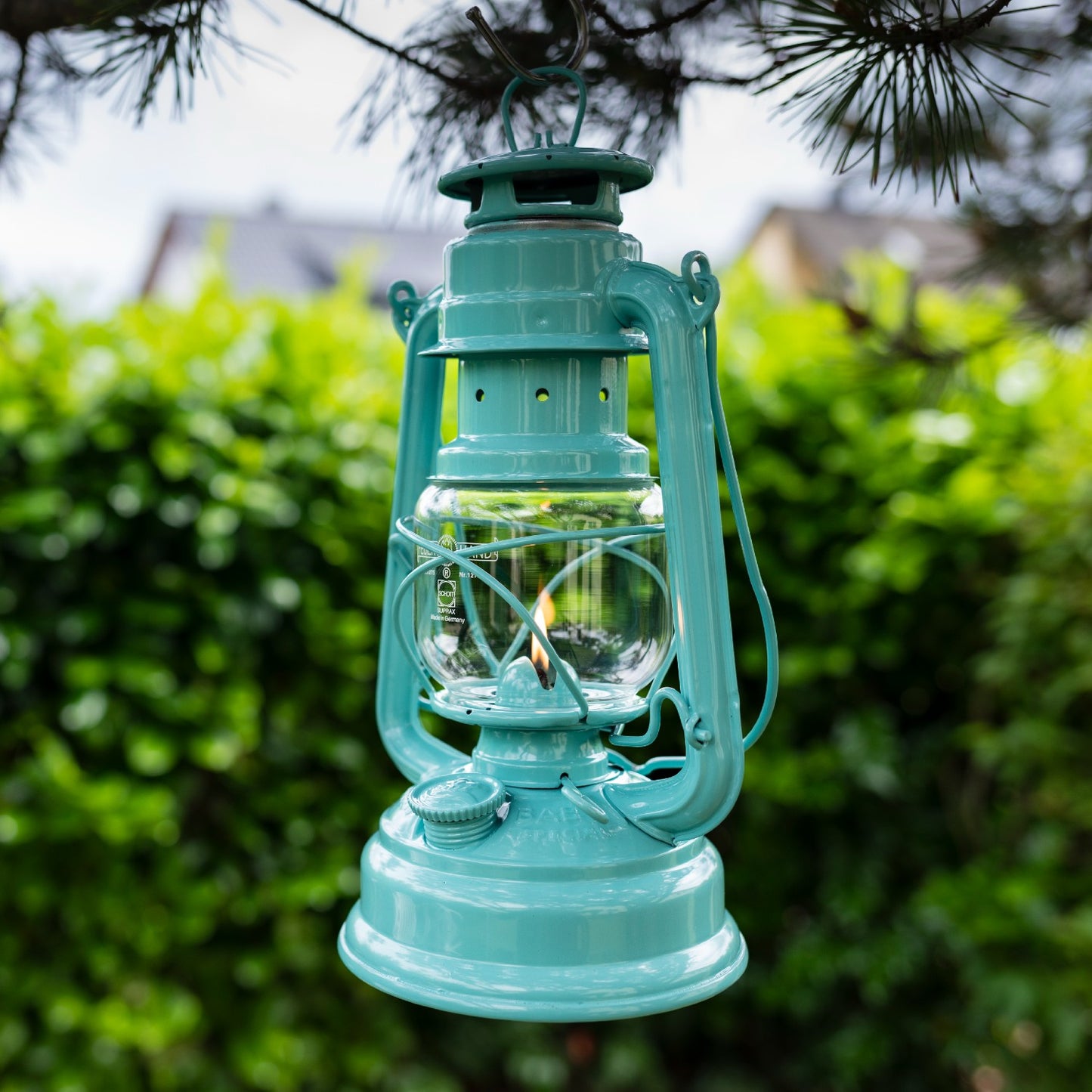Handcrafted turquoise Feuerhand Galvanized Oil Lantern with thermal heat-resistant SUPRAX globe softly glowing outdoors, hanging from a tree branch, ideal for Lamps & Lighting, Camping, and Emergency Supplies collections.