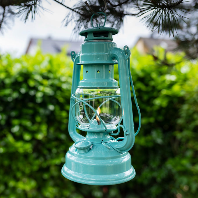 Handcrafted turquoise Feuerhand Galvanized Oil Lantern with thermal heat-resistant SUPRAX globe softly glowing outdoors, hanging from a tree branch, ideal for Lamps & Lighting, Camping, and Emergency Supplies collections.