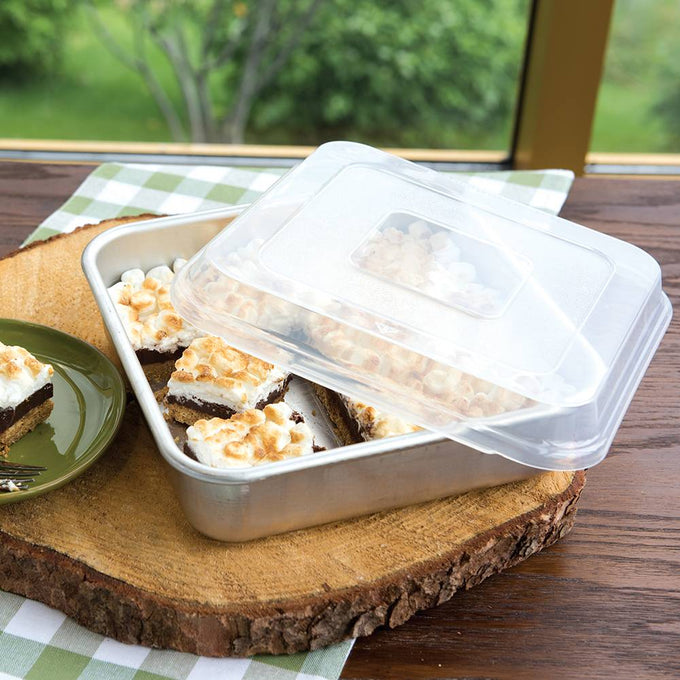 Square aluminum cake pan with encapsulated galvanized steel rim and clear plastic lid, holding toasted marshmallow dessert bars on a wooden slab, perfect for kitchen and baking supplies.
