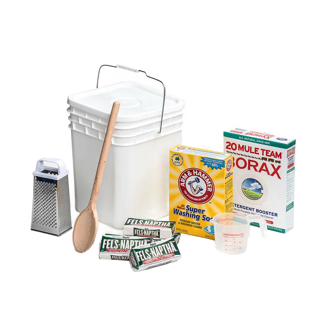 Plastic bucket with lid and metal handle alongside wooden spoon, metal grater, measuring cup, bar soaps, washing soda, and borax for making homemade laundry soap, handcrafted in the USA, suited for home and homesteading uses.