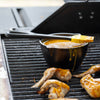 Black cast iron melting pot filled with sauce and topped with a silicone basting brush, placed on an outdoor grill alongside grilled chicken wings, ideal for Kitchen & Food and Cast Iron cookware collections.