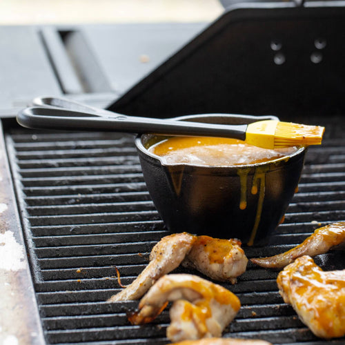 Black cast iron melting pot filled with sauce and topped with a silicone basting brush, placed on an outdoor grill alongside grilled chicken wings, ideal for Kitchen & Food and Cast Iron cookware collections.