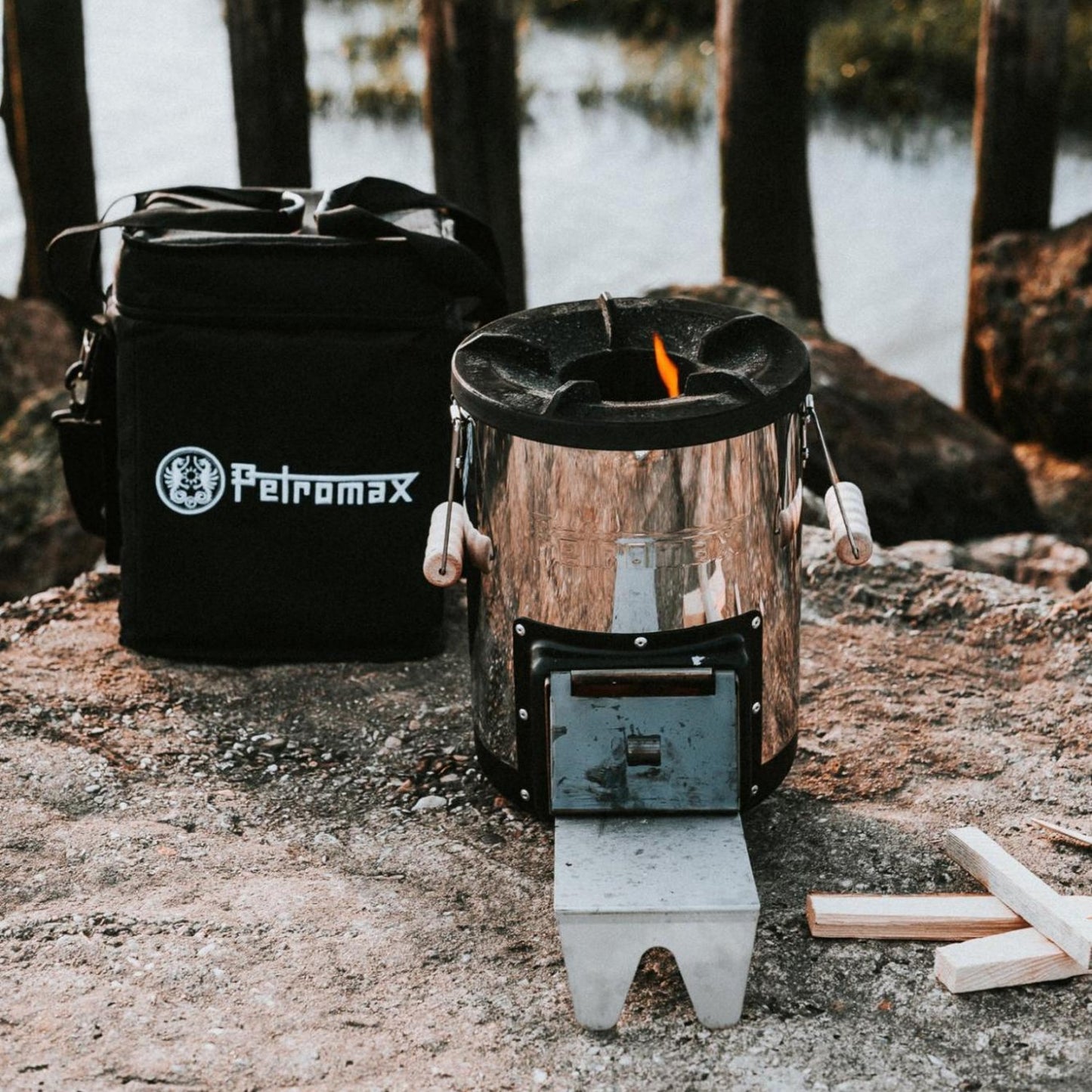 Compact metal rocket stove with flame burning, paired with a black durable transport bag featuring reinforced sides and a shoulder strap, designed for camping and farm use.