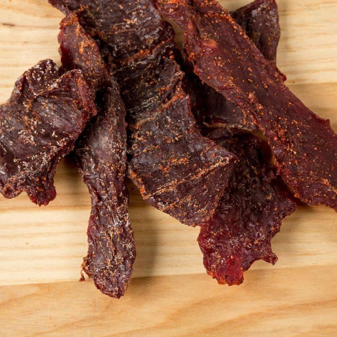 Close-up of dark, seasoned traditional beef jerky pieces on a wooden surface, showcasing a protein-rich, handcrafted snack made in USA for Kitchen & Food, Food, Snacks and Sweets, Meats, USA Made, Best Sellers, and Promo Eligible collections.