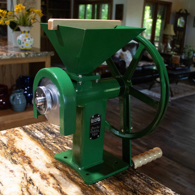 Green sturdy grain mill with large hand crank and wooden handle, featuring stainless steel burrs and a metal badge, suitable for homesteading, kitchen, and emergency supplies.