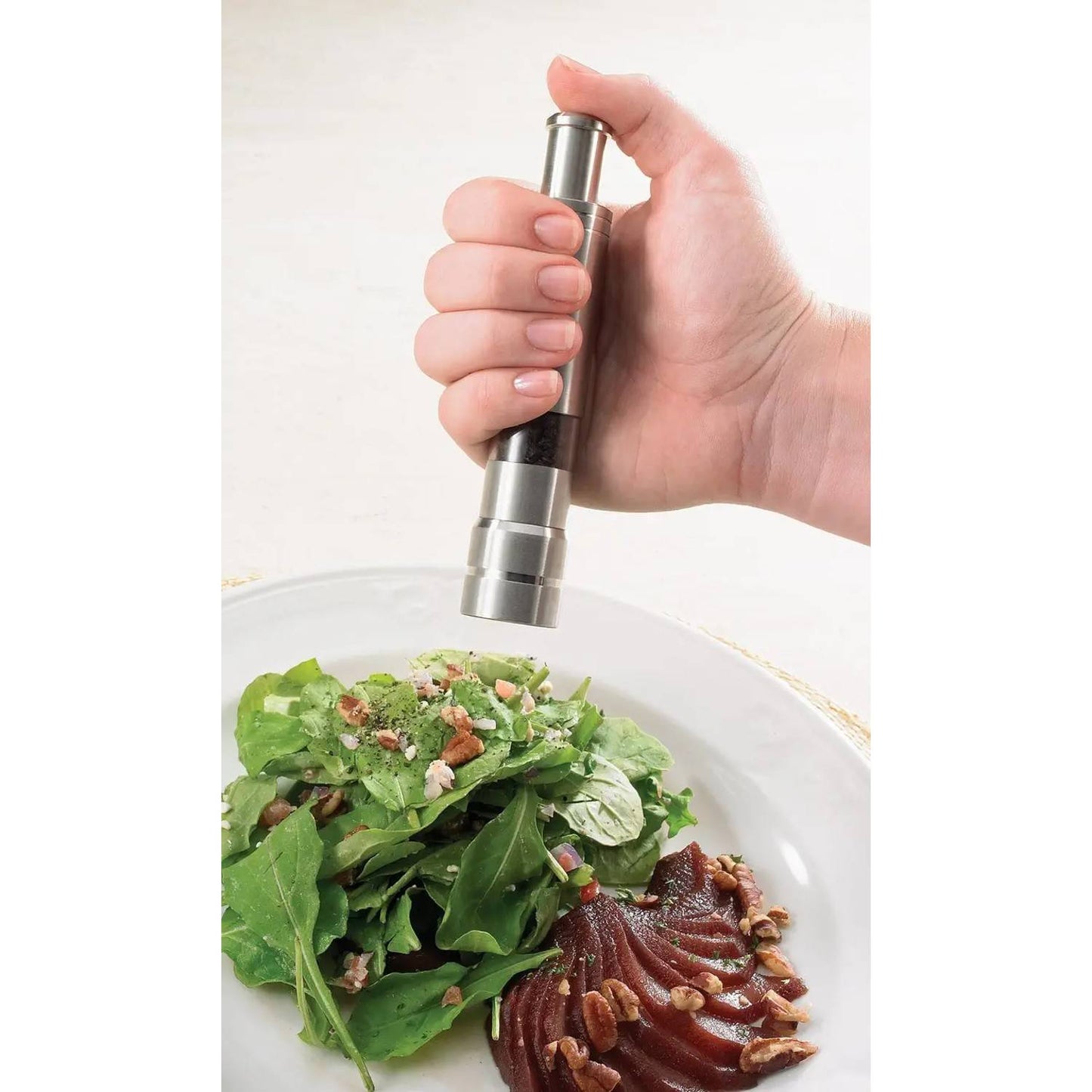 Hand holding a stainless steel one-handed pepper grinder dispensing freshly ground pepper over a salad with leafy greens and poached pear, featured in Kitchen & Food collections.