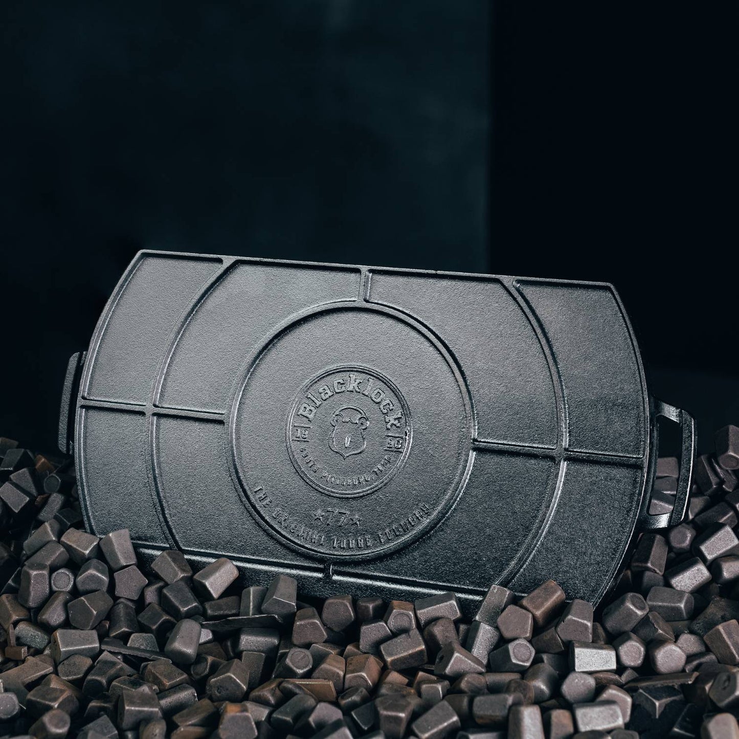 Top view of a handcrafted Lodge Blacklock cast iron double burner griddle with raised ridges, positioned on a bed of dark cast iron seasoning cubes, featured in Kitchen & Food, Cast Iron, USA Made, and Best Sellers collections.