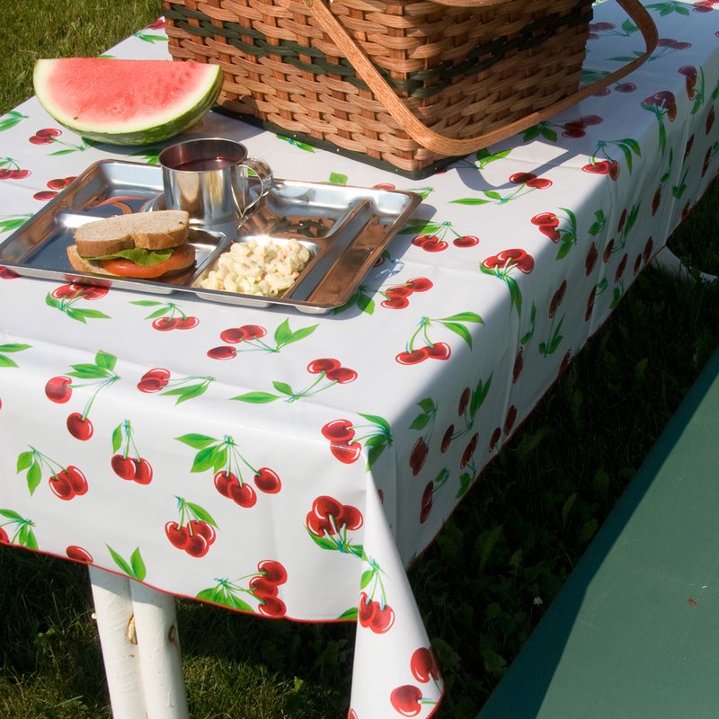 Outdoor picnic setup featuring a wipe-clean vintage vinyl tablecloth with bright red cherry patterns and red serged edges, shown with a picnic basket, sandwich, macaroni salad, and watermelon slice, ideal for Home & Decor, Kitchen & Food, and Kitchen Textiles collections.