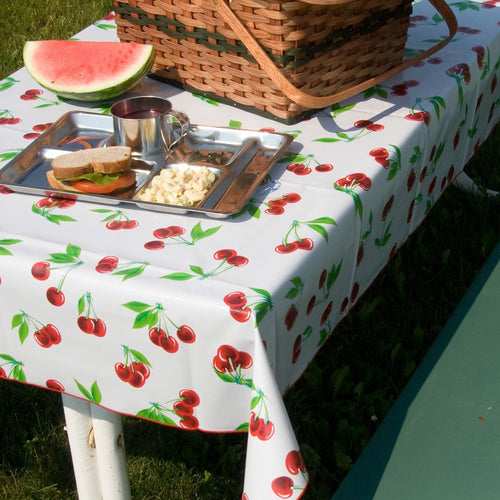 Outdoor picnic setup featuring a wipe-clean vintage vinyl tablecloth with bright red cherry patterns and red serged edges, shown with a picnic basket, sandwich, macaroni salad, and watermelon slice, ideal for Home & Decor, Kitchen & Food, and Kitchen Textiles collections.