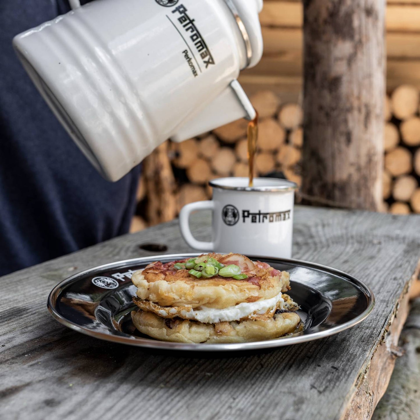 Black enamel plate with white rim and Petromax logo holding a breakfast sandwich topped with green onions, paired with a matching enamel mug and kettle pouring coffee, perfect for Farm, Garden & Tool, Kitchen & Food, Camping, Dinnerware, Best Sellers, and Promo Eligible collections.