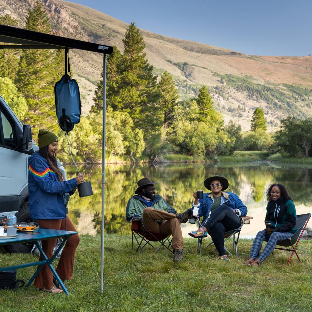 Group of four people camping by a lakeside with a gravity water filter hanging from a van awning, surrounded by nature in a peaceful outdoor setting suitable for camping and emergency supplies collections
