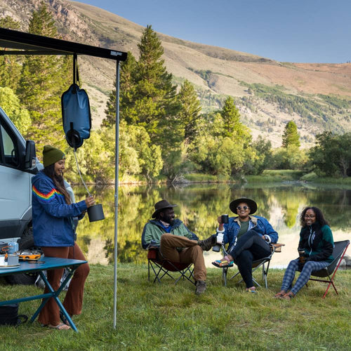 Group of four people camping by a lakeside with a gravity water filter hanging from a van awning, surrounded by nature in a peaceful outdoor setting suitable for camping and emergency supplies collections
