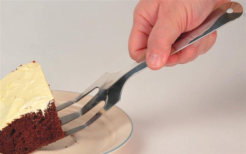 Hand holding stainless steel all-in-one kitchen tool spatula with angled, slotted, sharpened edge lifting a slice of chocolate cake with white frosting on a white plate, made in USA, ideal for Kitchen & Food, Storage and Serving, USA Made, and Best Sellers collections.