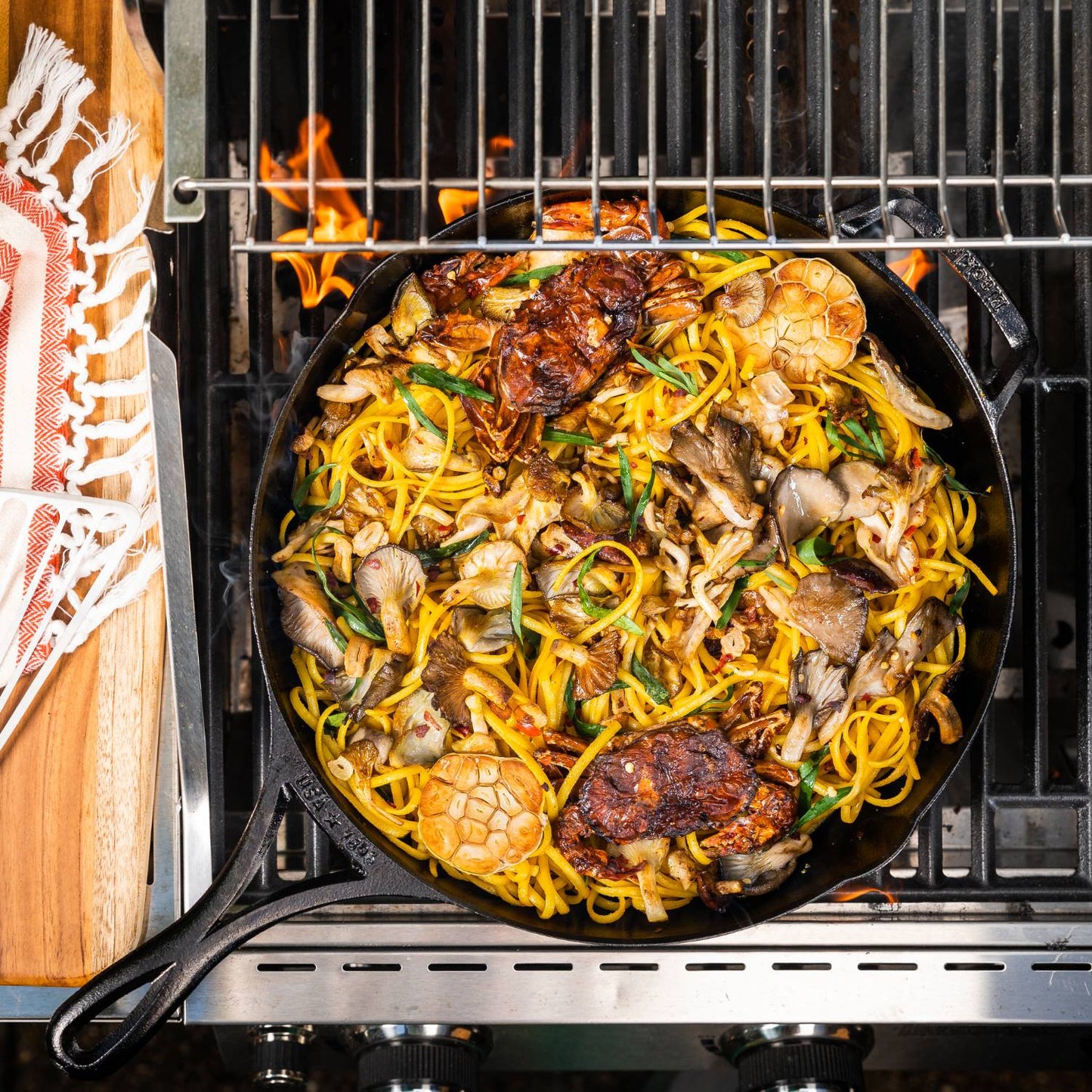 Handcrafted lightweight cast iron skillet cooking a vibrant stir-fry with noodles, mushrooms, herbs, and garlic heads on a grill, suited for Kitchen & Food, Cookware, and USA Made collections.