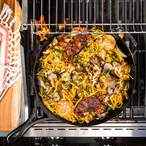 Handcrafted lightweight cast iron skillet cooking a vibrant stir-fry with noodles, mushrooms, herbs, and garlic heads on a grill, suited for Kitchen & Food, Cookware, and USA Made collections.