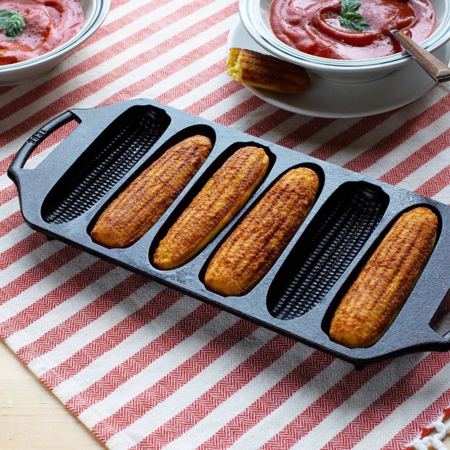 Lodge cast iron cornstick pan with six golden cornbread sticks, dual handles for safe handling, angled feet for oven stability, displayed on striped cloth beside bowls of tomato soup, ideal for Kitchen & Food, Cookware, Cast Iron, USA Made, Best Sellers, and Promo Eligible collections.