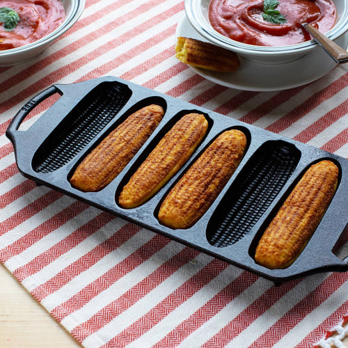 Lodge cast iron cornstick pan with six golden cornbread sticks, dual handles for safe handling, angled feet for oven stability, displayed on striped cloth beside bowls of tomato soup, ideal for Kitchen & Food, Cookware, Cast Iron, USA Made, Best Sellers, and Promo Eligible collections.