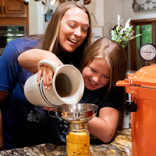 Mother and daughter happily pouring liquid from a handcrafted Heritage Blue Stripe Stoneware Pitcher through a funnel into a jar, in a cozy kitchen setting ideal for Kitchen & Food and USA Made collections.