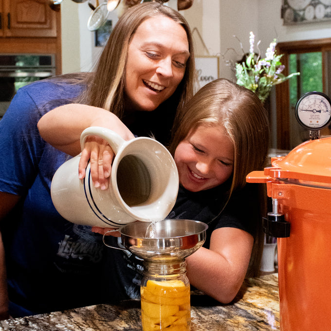 Mother and daughter happily pouring liquid from a handcrafted Heritage Blue Stripe Stoneware Pitcher through a funnel into a jar, in a cozy kitchen setting ideal for Kitchen & Food and USA Made collections.