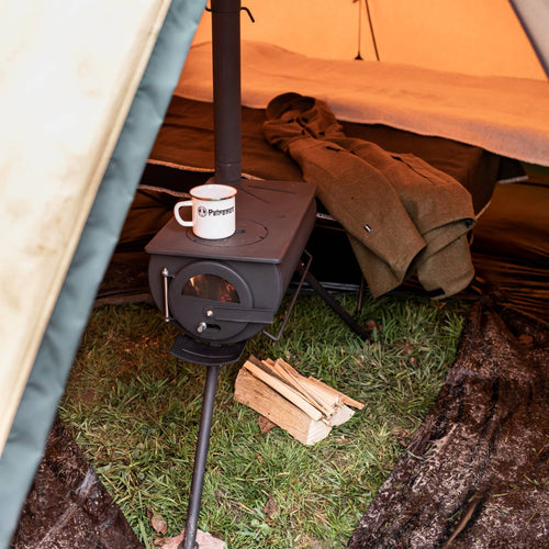 Compact black wood-burning tent heater with a mug on top, supported by foldable legs inside a camping tent, accompanied by firewood and outdoor gear, crafted for Farm, Garden & Tool and Camping collections.