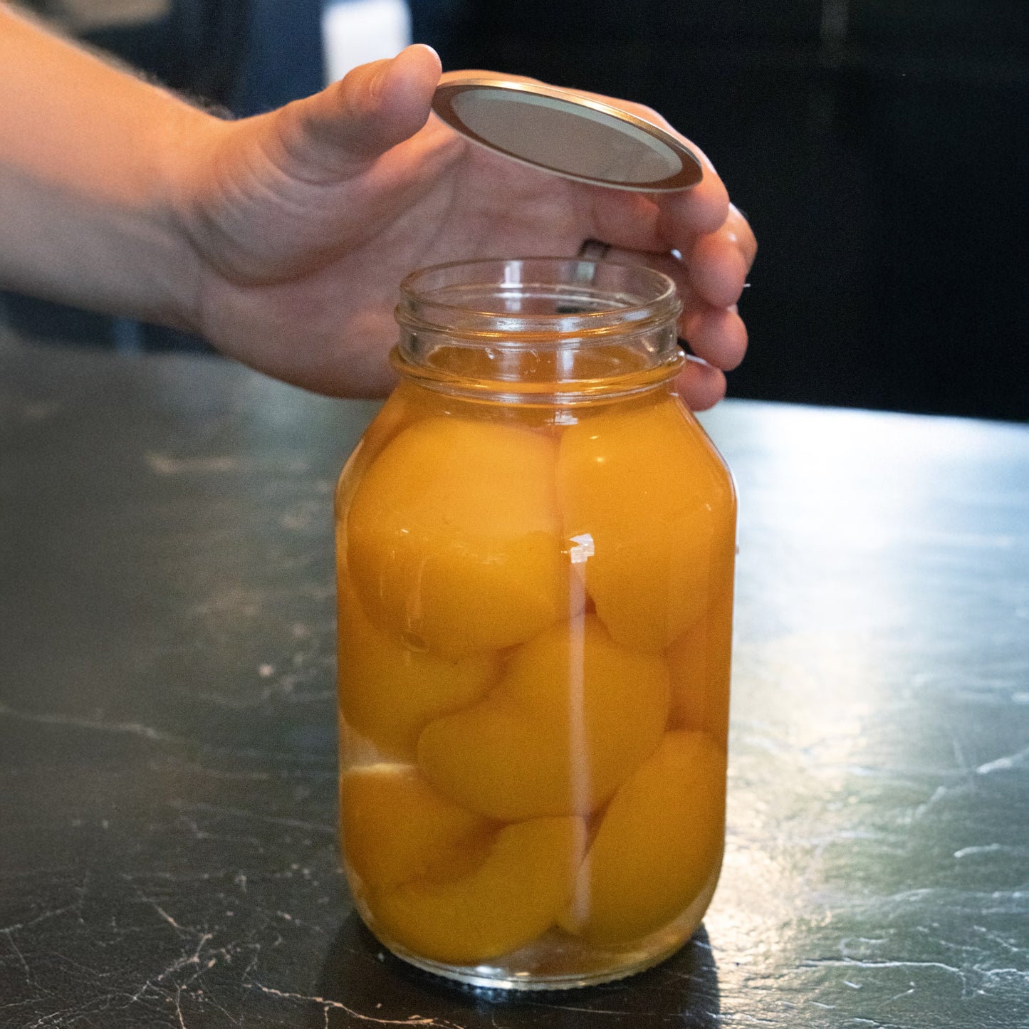Hand placing a lid with BPA-free sealing compound on a regular-mouth glass jar filled with preserved peaches, suitable for Kitchen & Food, Food Preservation, Canning, and Promo Eligible collections.