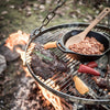Chromium-plated steel grate suspended by adjustable chains over an open campfire, grilling steak, peppers, and a cast iron skillet with beans, suited for outdoor camping and farm use collections.