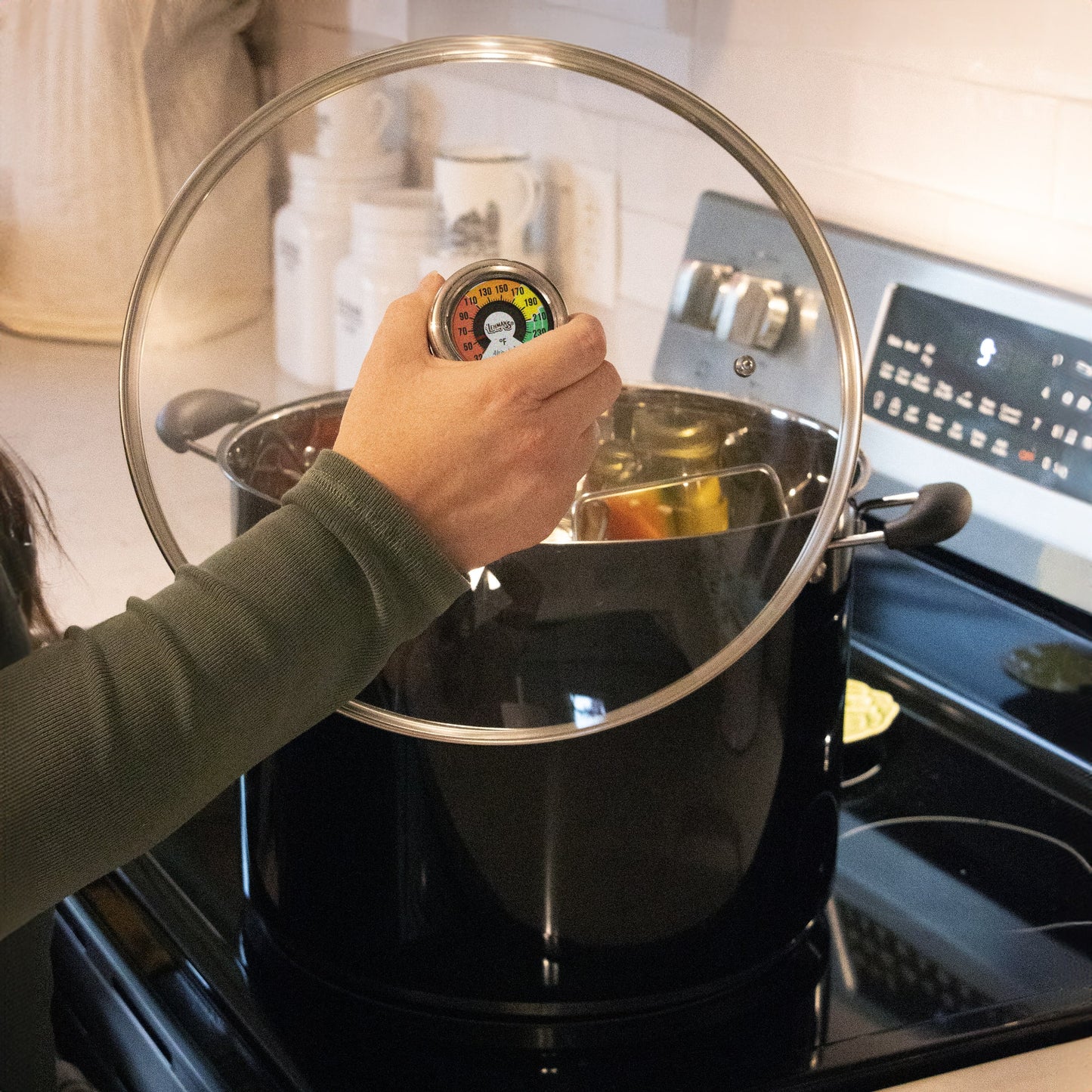 Hand adjusting built-in temperature gauge on a glass lid of a black 21.5-quart water bath canner heating on stove, part of Kitchen & Food, Food Preservation, and Canning collections.