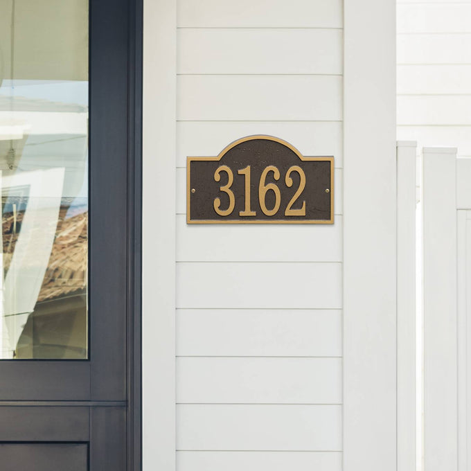 Handcrafted custom address plaque in bronze and gold with arched rectangle design, mounted on white siding near a glass-paneled door, featured in Home & Decor and USA Made collections.