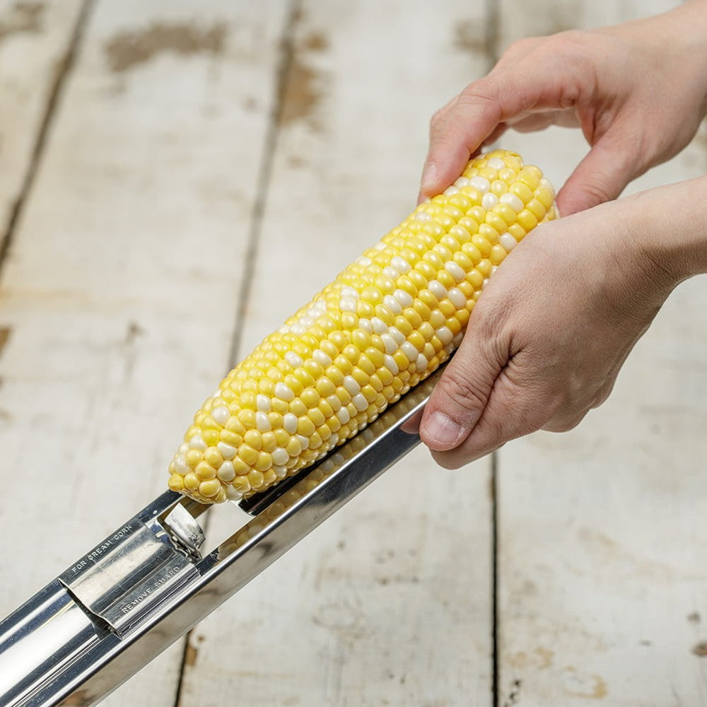 Person using a stainless steel corn cutter to easily remove kernels from a fresh ear of corn on a rustic wooden surface, suitable for Kitchen & Food and Food Prep collections.