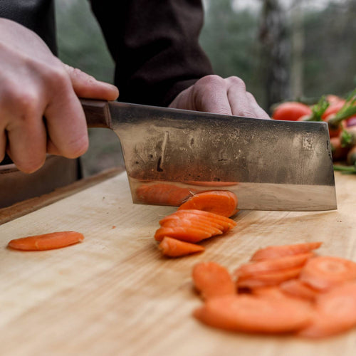 Handcrafted cleaver knife with a robust blade slicing fresh carrots on a wooden cutting board, ideal for kitchen food prep, camping, and farm tool collections.