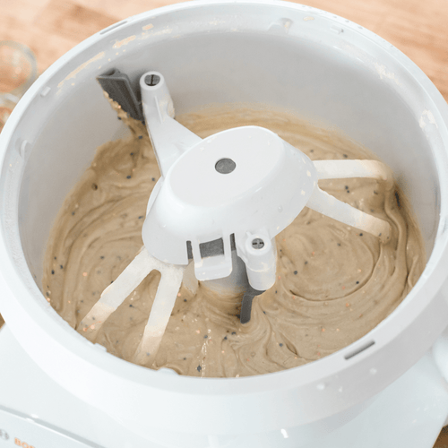 White bowl scraper attachment gently scraping thick batter from the sides and inner post of a mixer bowl, a handcrafted USA made kitchen gadget for baking and cooking helpers collections
