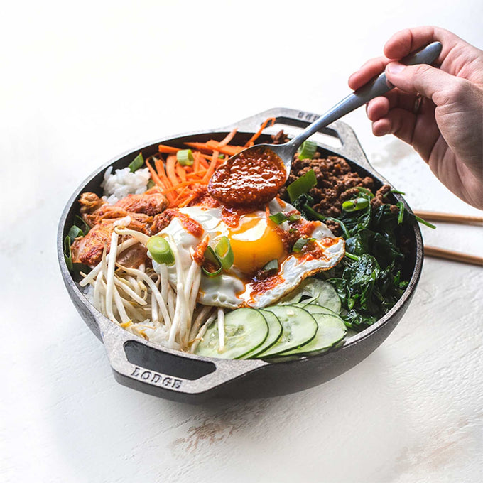 Handcrafted cast iron mini wok with sloped sides and loop handles, featuring a vibrant bibimbap dish, including fried egg, vegetables, and sauce, ideal for Kitchen & Food and Cookware collections.